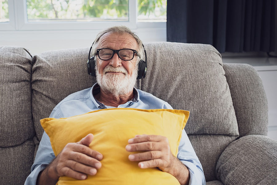 An elderly man sitting on a couch smiling while wearing headphones and holding a pillow