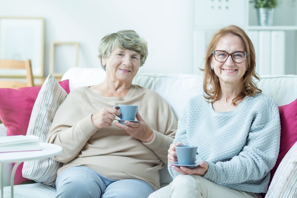 Aged smiling carer and sick older woman drinking tea together Planned social activities 7 days a week, such as card games or art lessons