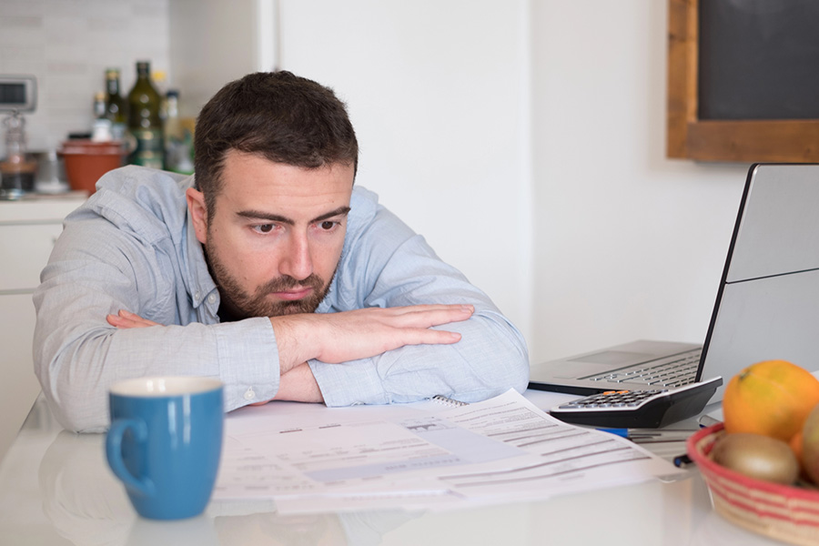 A man exhibiting caregiver stress signs as he looks exhausted while reviewing financial paperwork and bills at a kitchen table. A man exhibiting caregiver stress signs as he looks exhausted while reviewing financial paperwork and bills at a kitchen table.