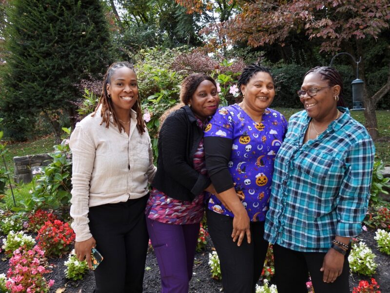 Caregivers smiling together in a garden, representing the supportive team environment of residential dementia care.