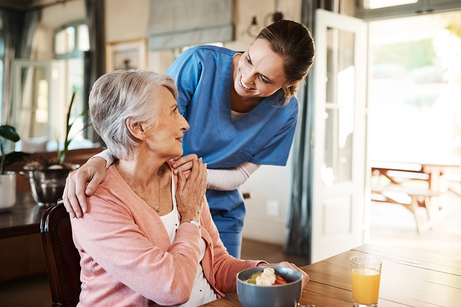 Caregiver smiling beside an elderly resident during her breakfast in a cozy memory care home near Sandy Spring, Maryland Caregiver smiling beside an elderly resident during her breakfast in a cozy memory care home near Sandy Spring, Maryland