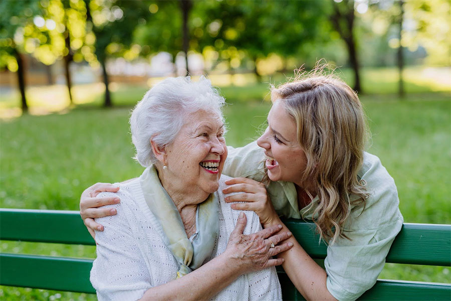 Elderly woman and younger caregiver smiling together outdoors, showing support during dementia progression.