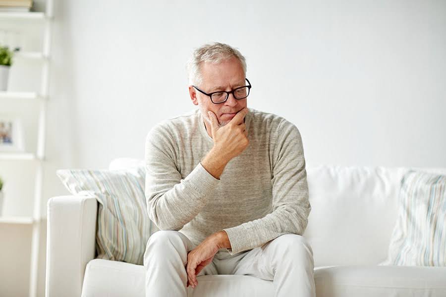 Older man sitting on a couch looking thoughtful, representing the question Why Can’t I Remember Anything Older man sitting on a couch looking thoughtful, representing the question Why Can’t I Remember Anything