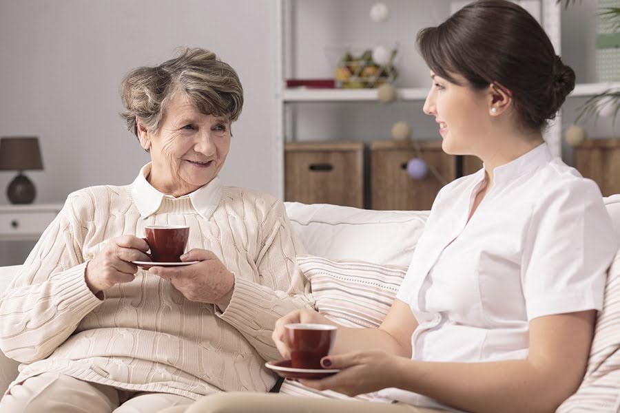 senior living communities caregiver chatting with an older woman on a couch while holding tea cups senior living communities caregiver chatting with an older woman on a couch while holding tea cups