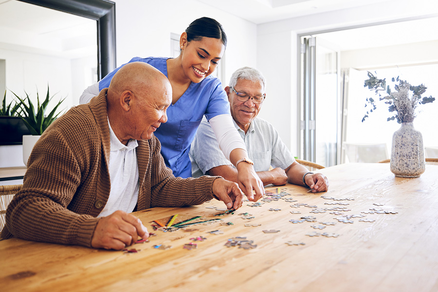 A caregiver providing support in a residential memory care home while two senior men work on a jigsaw puzzle at a wooden dining table