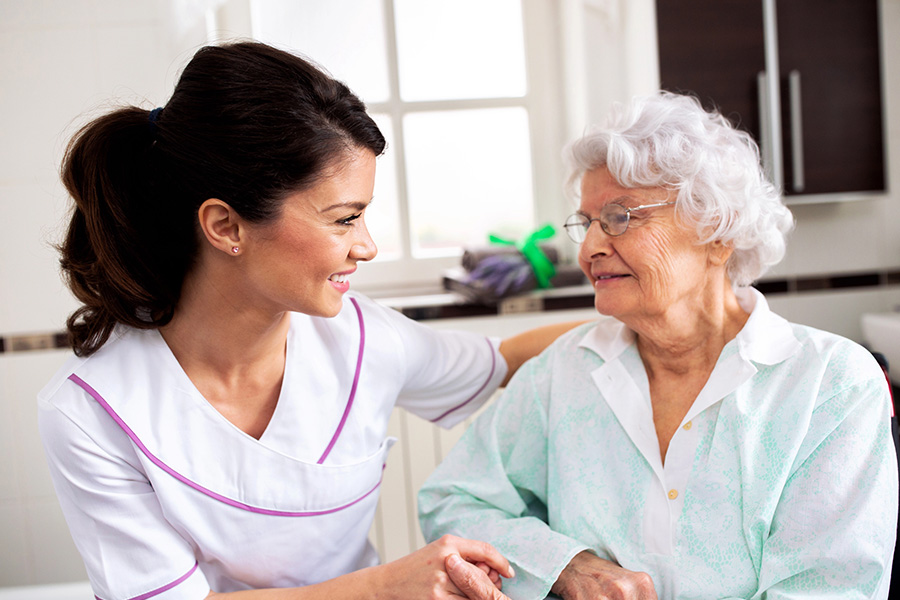 A smiling caregiver providing person centered dementia support while holding hands and comforting an elderly resident in a bright residential setting