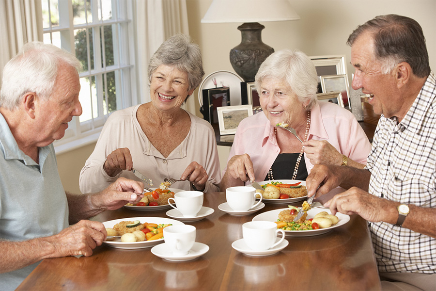 Four seniors smiling and enjoying a family-style dinner at a cozy Cedar Creek Memory Care home dining table.