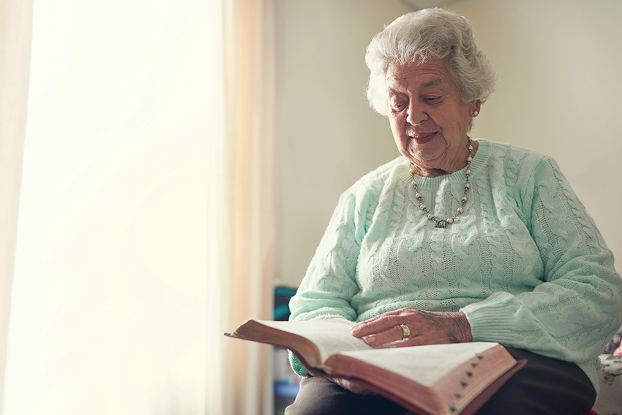 Senior woman quietly reading a book by a window, maintaining cognitive engagement and daily autonomy.